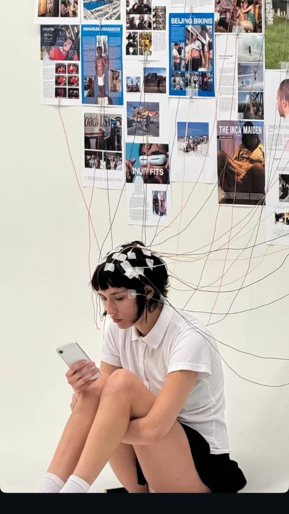 Person wired to a wall of random magazine clippings while scrolling a phone