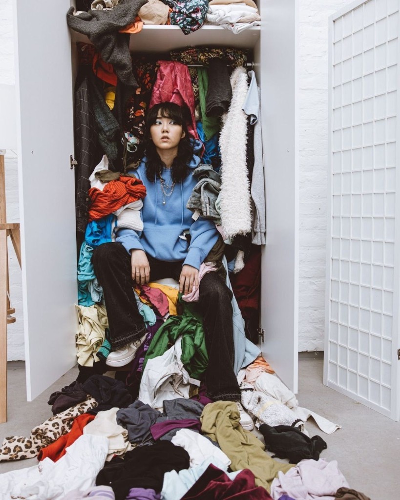 Person sitting in an overflowing open wardrobe surrounded by piled clothes