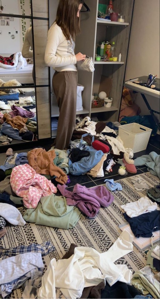 Person alone in a messy bedroom surrounded by clothes, getting dressed without help